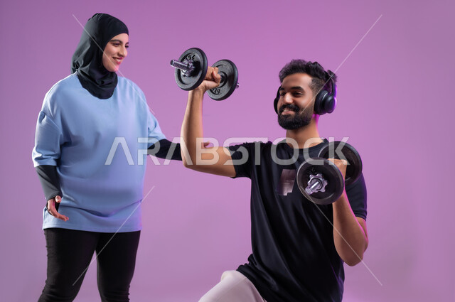نشاط بدني للمحافظة على الصحة واللياقة الجسدية، بورتريه لمدربة عربية خليجية سعودية ترتدي الزي الرياضي تقوم بتدريب رجل يرتدي سماعة الهيدفون يقوم برفع الأوزان الحديدية (الدمبلز)، مفهوم الرشاقة واللياقة البدنية، انشطة ورياضات لتقوية عضلات الجسم،  خلفية بنفسجية