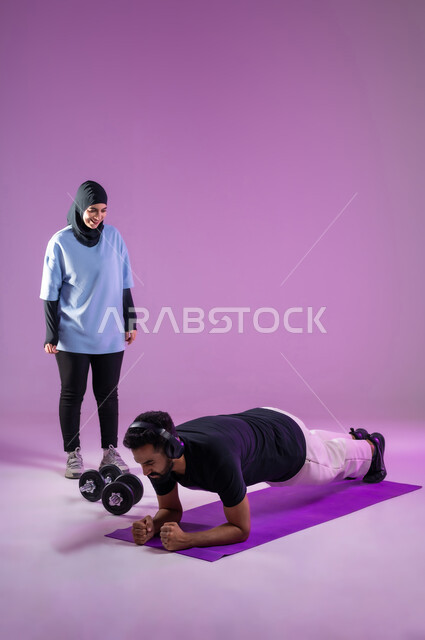 المحافظة على توازن الجسم بواسطة حركة البلانك، بورتريه لمدربة عربية خليجية سعودية محجبة ترتدي الزي الرياضي تقوم بتدريب رجل يرتدي سماعة الهيدفون ويقوم بممارسة تمارين الضغط, تقوية الأعصاب وعضلات الجسم، اسلوب حياة صحي، مفهوم الرشاقة واللياقة البدنية, خلفية بنفسجية