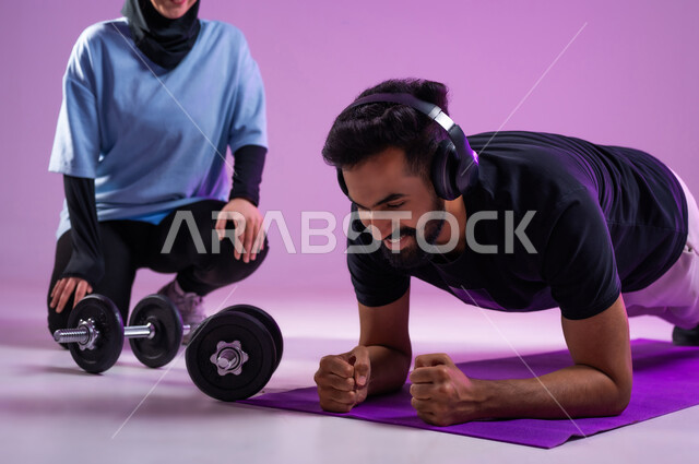 المحافظة على التوازن الجسدي, بورتريه لمدربة عربية خليجية سعودية محجبة ترتدي الزي الرياضي تقوم بتدريب رجل يرتدي سماعة الهيدفون ويقوم بممارسة تمارين الضغط, تقوية الأعصاب وعضلات الجسم، مفهوم الرشاقة واللياقة البدنية، انشطة ورياضات لتقوية العضلات, خلفية بنفسجية