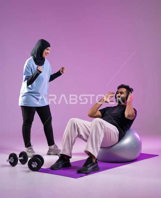 مفهوم الرشاقة واللياقة، بورتريه لمدربة عربية خليجية سعودية ترتدي الزي الرياضي ترفع يديها بايماءات الحماس والتشجيع، رجل يرتدي سماعات الهيدفون يمارس تمارين الاحماء باستخدام كرة البيلاتس، المحافظة على التوازن الجسدي، ادوات رفع الاثقال، خلفية بنفسجية