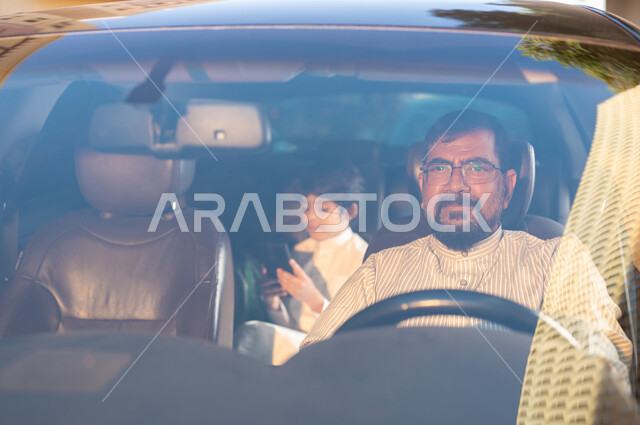 Mobile programs and applications, the concept of driving and delivery, Saudi professions and jobs, recruiting workers from abroad, a smiling Pakistani driver wearing medical glasses transporting a Saudi Gulf Arab child holding a mobile phone in his hand, using modern techniques and technology