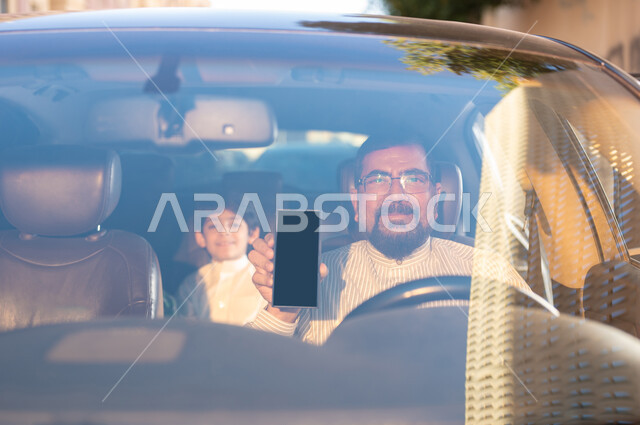 استخدام التقنيات والوسائل الحديثة، مفهوم القيادة والتوصيل، مهن ووظائف سعودية، استقدام العمالة من الخارج، سائق باكستاني مبتسم يرتدي النظارة الطبية يمسك بيده الهاتف المحمول بشاشة سوداء فارغة يقوم بنقل طفل عربي خليجي سعودي، برامج وتطبيقات الجوال