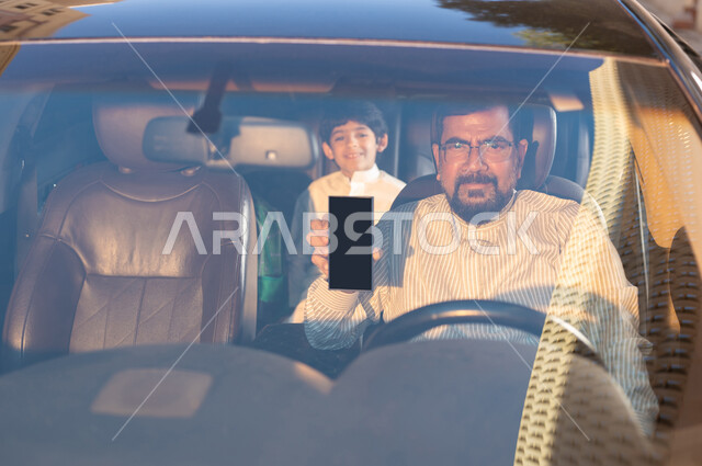 استخدام التقنيات والوسائل الحديثة، مفهوم القيادة والتوصيل، مهن ووظائف سعودية، استقدام العمالة من الخارج، سائق باكستاني مبتسم يرتدي النظارة الطبية يمسك بيده الهاتف المحمول بشاشة سوداء فارغة يقوم بنقل طفل عربي خليجي سعودي، برامج وتطبيقات الجوال