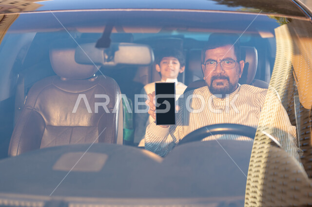 استخدام التقنيات والوسائل الحديثة، مفهوم القيادة والتوصيل، مهن ووظائف سعودية، استقدام العمالة من الخارج، سائق باكستاني مبتسم يرتدي النظارة الطبية يمسك بيده الهاتف المحمول بشاشة سوداء فارغة يقوم بنقل طفل عربي خليجي سعودي، برامج وتطبيقات الجوال