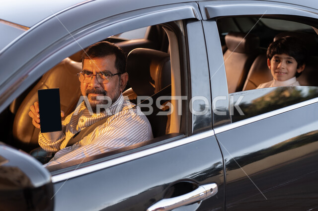 Using modern technologies and means, the concept of driving and delivery, Saudi professions and jobs, recruiting workers from abroad, a smiling Pakistani driver wearing medical glasses holding a mobile phone with a blank black screen in his hand transporting a Saudi Gulf Arab child, mobile programs and applications