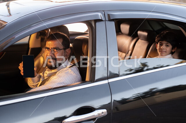 استخدام التقنيات والوسائل الحديثة، مفهوم القيادة والتوصيل، مهن ووظائف سعودية، استقدام العمالة من الخارج، سائق باكستاني مبتسم يرتدي النظارة الطبية يمسك بيده الهاتف المحمول بشاشة سوداء فارغة يقوم بنقل طفل عربي خليجي سعودي، برامج وتطبيقات الجوال