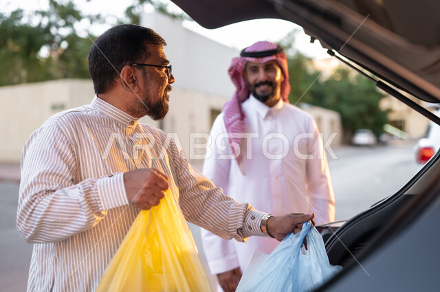 اقتناء المستلزمات المنزلية، استقطاب العمالة من الخارج، شاب سعودي خليجي يرتدي الشماغ والثوب التقليدي يقف مع رجل باكستاني يمسك بيده اكياس الحوائج يقوم بوضعها في السيارة يتناقشان بإيماءات السرور