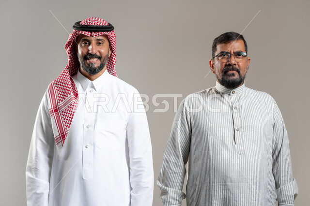 The concept of elegance and masculinity, attention to external appearance, attracting workers from abroad, a close-up portrait of a Saudi Gulf Arab man wearing a shemagh and a traditional thobe, standing with a worker wearing a Pakistani thobe, looking at the camera with gestures of joy and pleasure, gray background