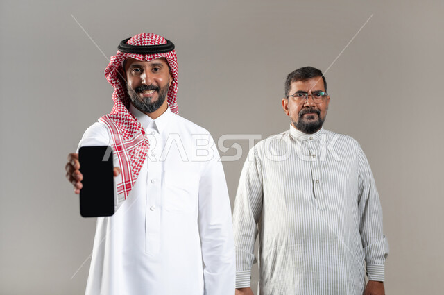 التقنيات الحديثة والتكنولوجيا، النظر إلى الكاميرا بإيماءات السرور، بورتريه مقرب لرجل عربي سعودي خليجي يرتدي الشماغ والثوب التقليدي يعرض جهاز الهاتف المحمول بشاشة سوداء فارغة يقف مع عامل يرتدي الثوب الباكستاني، خلفية رمادية