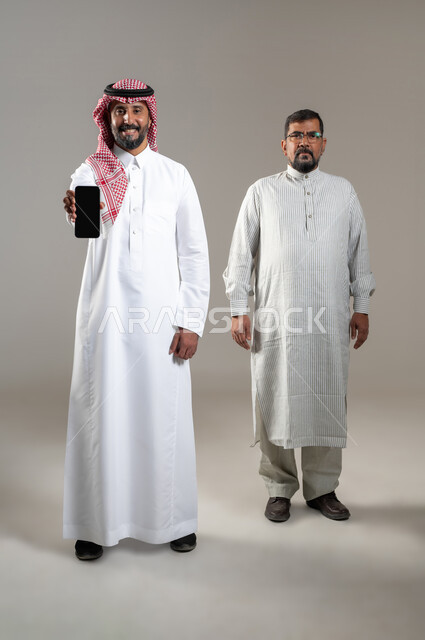 Technology and modern techniques, looking at the camera with gestures of pleasure, portrait of a Saudi Gulf Arab man wearing a shemagh and traditional thobe, showing a mobile phone device with a blank black screen, standing with a worker wearing a Pakistani thobe, full-length body portrait, gray background