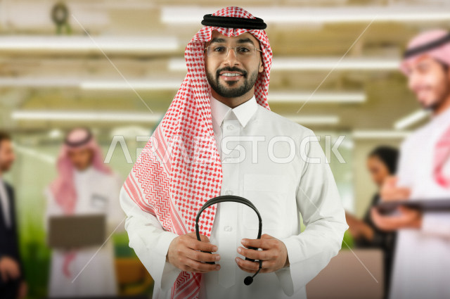 شاب موظف خليجي سعودي  في بيئة العمل يحمل بيده سماعة رأس صوتية  ، العمل بالدعم الفني ، الكول سنتر ، خلفية مكان العمل وزملاء العمل