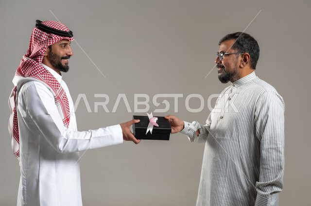 مشاعر الحب بين الاصدقاء، بورتريه مقرب لشاب خليجي سعودي يقوم بتقديم هدية لصديقه يرتدي الثوب الباكستاني، المشاركه في الأعياد والإحتفالات، ايماءات السعادة والدهشة والسرور، خلفية رمادية