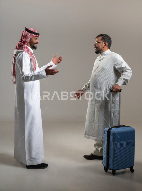 نقل حقائب الأمتعة، استقدام العمالة من الخارج، بورتريه لرجل عربي سعودي يرتدي الشماغ والثوب التقليدي مع شاب يرتدي الثوب الباكستاني يمسك حقيبة السفر يتحادثان بإيماءات السرور والسعادة، صورة الجسم بالطول الكامل، خلفية رمادية