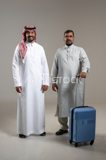 نقل حقائب الأمتعة، استقدام العمالة من الخارج، بورتريه لرجل عربي سعودي يرتدي الشماغ والثوب التقليدي مع شاب يرتدي الثوب الباكستاني يمسك حقيبة السفر ينظران الى الكاميرا بإيماءات السرور والسعادة، صورة الجسم بالطول الكامل، خلفية رمادية