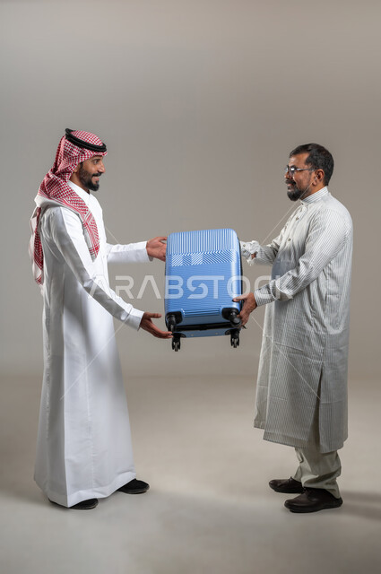 المساعدة في نقل حقائب الأمتعة، استقدام العمالة من الخارج، بورتريه لرجل عربي سعودي يرتدي الشماغ والثوب التقليدي يعطي حقيبة السفر لشاب يرتدي الثوب الباكستاني، إيماءات السرور والسعادة، صورة الجسم بالطول الكامل، خلفية رمادية