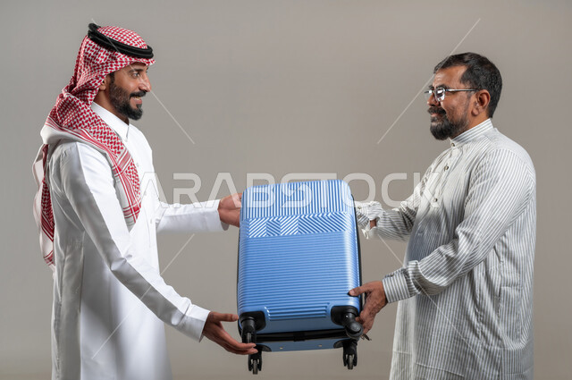 المساعدة في نقل حقائب الأمتعة، استقدام العمالة من الخارج، بورتريه مقرب لرجل عربي سعودي يرتدي الشماغ والثوب التقليدي يعطي حقيبة السفر لشاب يرتدي الثوب الباكستاني، إيماءات السرور والسعادة، خلفية رمادية