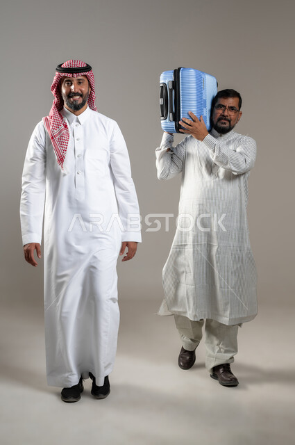 المساعدة في نقل حقائب الأمتعة، استقدام العمالة من الخارج، بورتريه لرجل عربي سعودي يرتدي الشماغ والثوب التقليدي مع شاب يرتدي الثوب الباكستاني يحمل حقيبة السفر ينظران الى الكاميرا بإيماءات السرور، تعابير السير والمشي، صورة الجسم بالطول الكامل، خلفية رمادية