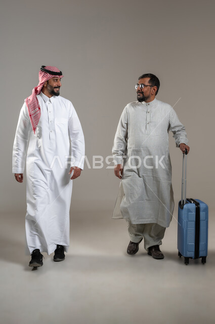 المساعدة في نقل حقائب الأمتعة، استقدام العمالة من الخارج، بورتريه لرجل عربي سعودي يرتدي الشماغ والثوب التقليدي مع شاب يرتدي الثوب الباكستاني يمسك حقيبة السفر يتحادثان بإيماءات السرور، تعابير السير والمشي، صورة الجسم بالطول الكامل، خلفية رمادية