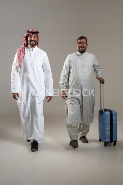 المساعدة في نقل حقائب الأمتعة، استقدام العمالة من الخارج، بورتريه لرجل عربي سعودي يرتدي الشماغ والثوب التقليدي مع شاب يرتدي الثوب الباكستاني يمسك حقيبة السفر ينظران الى الكاميرا بإيماءات السرور، تعابير السير والمشي، صورة الجسم بالطول الكامل، خلفية رمادية