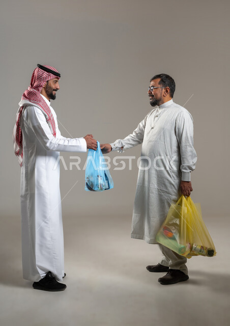 مفهوم استقدام العمالة من الخارج، بورتريه لرجل يرتدي الثوب الباكستاني يأخذ اكياس الحوائج والبضاعة من شاب سعودي يرتدي الشماغ والثوب التقليدي، صورة الجسم بالطول الكامل، خلفية رمادية