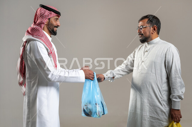 مفهوم استقدام العمالة من الخارج، بورتريه مقرب لرجل يرتدي الثوب الباكستاني يأخذ اكياس الحوائج والبضاعة من شاب سعودي يرتدي الشماغ والثوب التقليدي، شراء الاحتياجات و المستلزمات المنزلية للمستهلك، خلفية رمادية