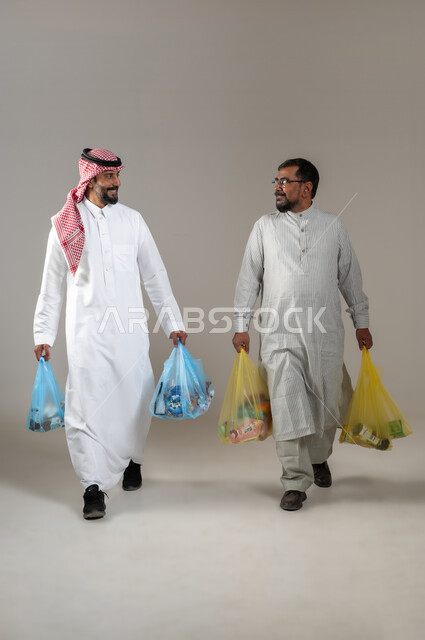 The concept of recruiting workers from abroad, a portrait of a man wearing a Pakistani thobe, holding bags of necessities and goods in his hand, talking with a young Saudi man wearing a shemagh and traditional thobe, with gestures of pleasure, walking and walking expressions, full-length body portrait, gray background