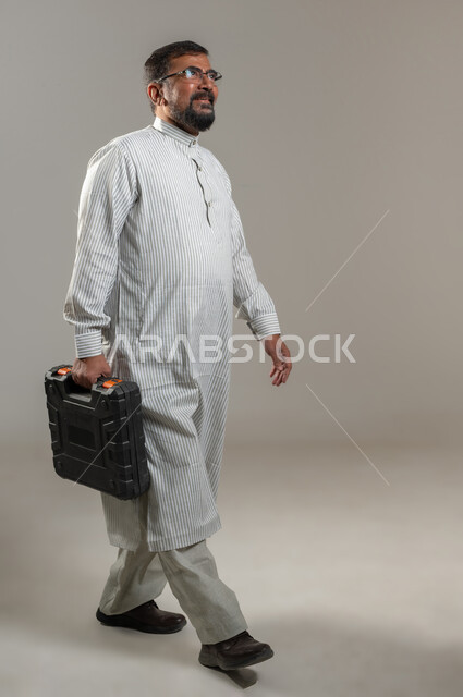 استقدام العمالة من الخارج، تصليح و صيانة السيارات، بورتريه لرجل يرتدي الثوب الباكستاني يحمل حقيبة المعدات والتجهيزات يمشي بتعابير الجدية والثقة بالنفس، صورة الجسم بالطول الكامل، خلفية رمادية