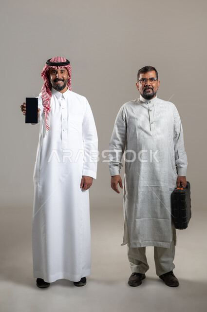 Car maintenance and repair, looking at the camera with pleased gestures, portrait of a Saudi Gulf Arab man wearing a shemagh and traditional thobe displaying a mobile phone device with a blank black screen, a worker wearing a Pakistani thobe holding an equipment bag, full-length body portrait, gray background