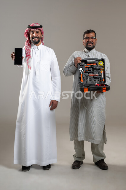 Car maintenance and repair, looking at the camera with pleased gestures, portrait of a Saudi Gulf Arab man wearing a shemagh and traditional thobe showing a mobile phone device with a blank black screen, a worker wearing a Pakistani thobe showing an equipment bag, full length body, gray background