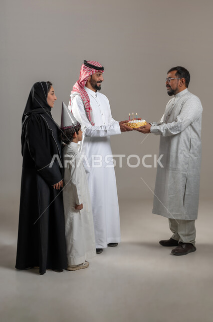 قضاء وقت ممتع مع الأصدقاء، مناسبات وأعياد عائلية سعيدة، مفهوم الترابط الأسري، إيماءات السعادة والمفاجئة، بورتريه مقرب لعائلة عربية خليجية سعودية تحمل قالب الكيك تقوم بتقديمه لرجل يرتدي الثوب الباكستاني والنظارة الطبية، صورة الجسم بالطول الكامل، خلفية رمادية