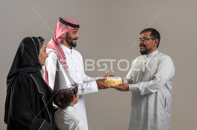 قضاء وقت ممتع مع الأصدقاء، مناسبات وأعياد عائلية سعيدة، مفهوم الترابط الأسري، إيماءات السعادة والمفاجئة والفرح، بورتريه مقرب لعائلة عربية خليجية سعودية تحمل قالب الكيك تقوم بتقديمه لرجل يرتدي الثوب الباكستاني و النظارة الطبية، خلفية رمادية