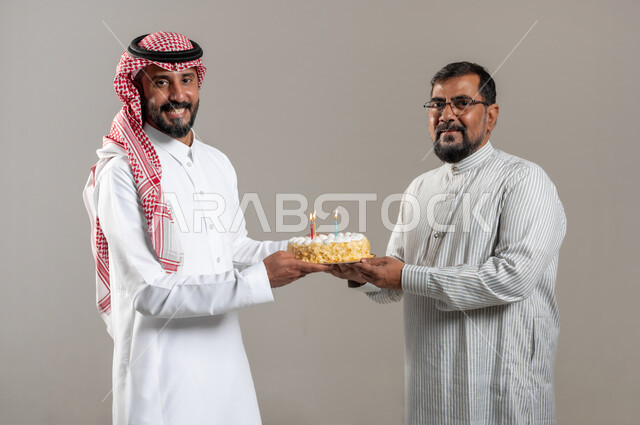 أعياد ومناسبات عائلية سعيدة، النظر الى الكاميرا بإيماءات السعادة والمفاجئة والفرح، بورتريه مقرب لرجل عربي خليجي سعودي يرتدي الشماغ والثوب التقليدي يقوم بتقديم قالب الكيك لصديقه، مفهوم الحب بين الأخوة، خلفية رمادية