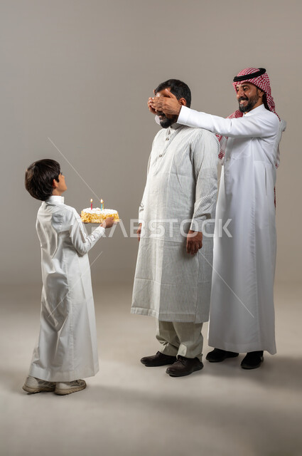إيماءات وجه ويدين تدل على الدهشة والمفاجئة والفرح، ولد عربي خليجي سعودي يقوم بتقديم قالب الكيك لوالده، بورتريه لرجل عربي خليجي سعودي يغمض عيني صديقه، أعياد ومناسبات عائلية سعيدة، صورة الجسم بالطول الكامل، خلفية رمادية