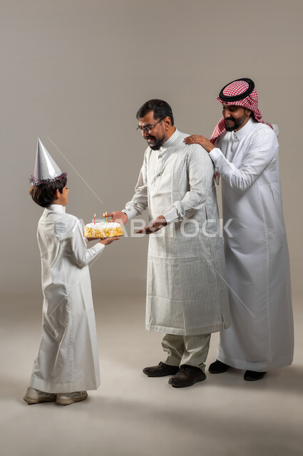إيماءات وجه ويدين تدل على الدهشة والمفاجئة والفرح، ولد عربي خليجي سعودي يقوم بتقديم قالب الكيك لوالده، بورتريه لرجل سعودي وشاب يرتدي الثوب الباكستاني يحتفلان بعيد الميلاد، أعياد ومناسبات عائلية سعيدة، صورة الجسم بالطول الكامل، خلفية رمادية