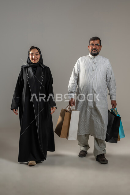 متعة التسوق واقتناء المستلزمات والهدايا، سيدة عربية خليجية سعودية محجبة ترتدي العباءة السوداء تستمتع بالتجول في المولات التجارية، رجل يرتدي الثوب الباكستاني يحمل بيده أكياس ورقية ملونة ينظر الى الكاميرا بإيماءات السرور، صورة الجسم بالطول الكامل, خلفية رمادية