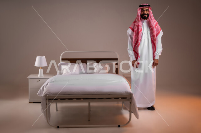 الإهتمام بالمظهر الخارجي، مفهوم الاناقة والرجولة، النظر إلى الكاميرا بايماءات الثقة بالنفس، تعابير السرور والسعادة، بورتريه لرجل عربي خليجي سعودي يرتدي الثوب التقليدي يقف بجانب السرير، صورة لكامل الجسم، خلفية ملونة