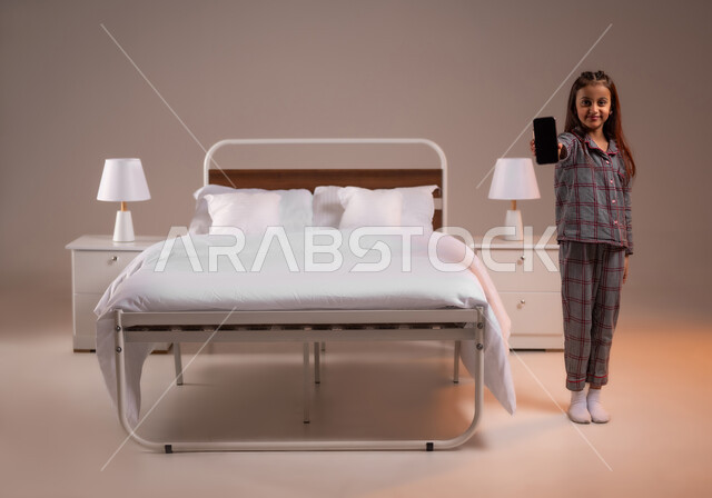 Development of technology and modern technical devices, displaying a blank black screen, looking at the camera with gestures of happiness, portrait of a smiling little Saudi Arabian Gulf girl wearing pajamas holding a mobile phone in her hand, full body image, colored background