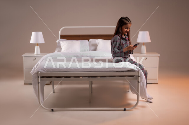 التواصل مع العائلة والاصدقاء عبر مواقع التواصل الاجتماعي، ايماءات الاندماج والتركيز، بورتريه لفتاه صغيرة عربيه سعوديه خليجيه تجلس على سرير مريح تقوم باستخدام الهاتف المحمول، خلفية ملونة