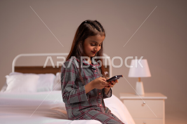 ايماءات الاندماج والتركيز، التواصل مع العائلة والاصدقاء عبر مواقع التواصل الاجتماعي، بورتريه لفتاه صغيرة عربيه سعوديه خليجيه تجلس على سرير مريح تقوم باستخدام الهاتف المحمول، خلفية ملونة