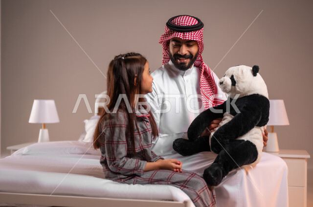 تقوية رابط الأبوة بين الأب وطفلته، تحمل المسؤولية والعناية بالابناء، قضاء أوقات ممتعة مع الاولاد، ممارسة الأنشطة الترفيهية المسلية، بورتريه لرجل عربي خليجي سعودي يقوم باللعب بدمية الدب مع ابنته، خلفية ملونة