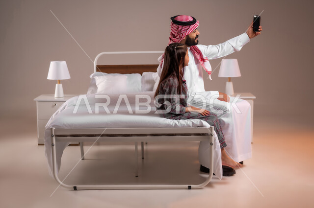 رعاية الأب لأطفاله والعناية بهم، قضاء أوقات ممتعة مع الابناء، مفهوم الأبوة والطفولة، إجراء مكالمة فيديو، بورتريه لرجل عربي خليجي سعودي يجلس على السرير برفقة ابنته يقوم بإلتقاط صورة سيلفي تذكارية بإستخدام الهاتف المحمول، الحب والحنان الاسري، خلفية ملونة