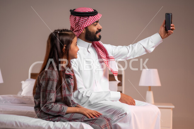 قضاء أوقات ممتعة مع الابناء، مفهوم الأبوة والطفولة، إجراء مكالمة فيديو، بورتريه لرجل عربي خليجي سعودي يجلس على السرير برفقة ابنته يقوم بإلتقاط صورة سيلفي تذكارية بإستخدام الهاتف المحمول، رعاية الأب لأطفاله والعناية بهم، الحب والحنان الاسري، خلفية ملونة