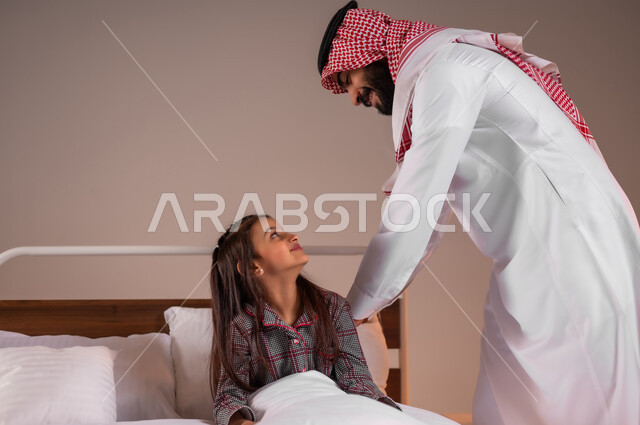 مفهوم العناية والاهتمام بالأبناء، اتباع أسلوب حياة صحي، الاسترخاء والاستمتاع بالأجواء الهادئة، قضاء اوقات عائلية سعيدة، بورتريه لرجل عربي خليجي سعودي يقوم بالاطمئنان على ابنته قبل النوم، خلفية ملونة