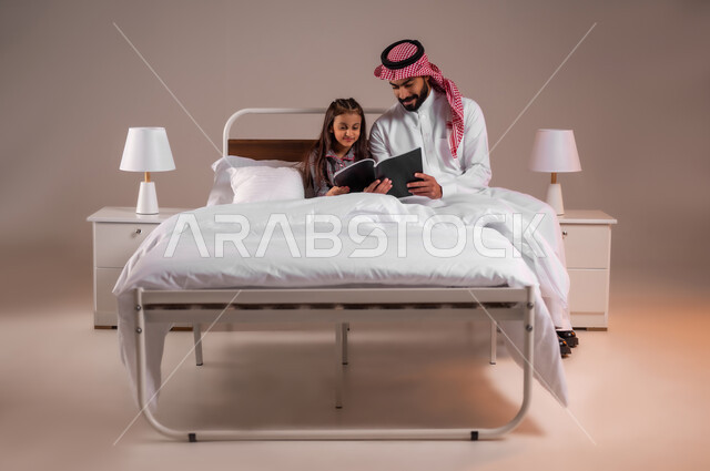 مفهوم التربيه القائمه على الحب والحنان، قضاء وقت ممتع مع الابنه، المشاركه بقراءه القصص المفيده مع الأبناء قبل النوم، بورتريه لرجل عربي خليجي سعودي يرتدي الثوب التقليدي والشماغ يجلس على السرير برفقه ابنته يقرأ الروايات المسليه، خلفية ملونة