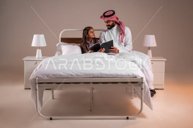 مفهوم التربيه القائمه على الحب والحنان، المشاركه بقراءه القصص المفيده مع الأبناء قبل النوم، قضاء وقت ممتع مع الابنه، بورتريه لرجل عربي خليجي سعودي يرتدي الثوب التقليدي والشماغ يجلس على السرير برفقه ابنته يقرأ الروايات المسليه، خلفية ملونة