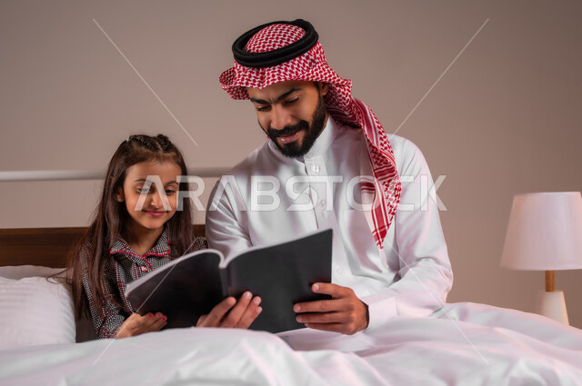 المشاركه بقراءه القصص المفيده مع الأبناء قبل النوم، قضاء وقت ممتع مع الابنه، مفهوم التربيه القائمه على الحب والحنان، بورتريه لرجل عربي خليجي سعودي يرتدي الثوب التقليدي والشماغ يجلس على السرير برفقه ابنته يقرأ الروايات المسليه، خلفية ملونة