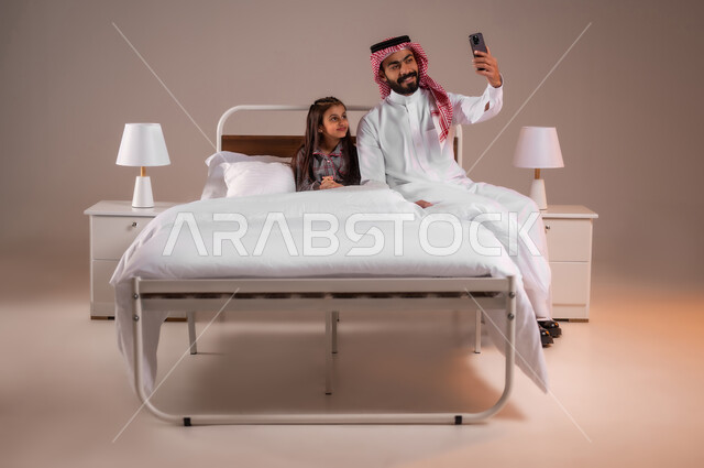 الحب والحنان الاسري، رعاية الأب لأطفاله والعناية بهم، قضاء أوقات ممتعة مع الابناء، مفهوم الأبوة والطفولة، إجراء مكالمة فيديو، بورتريه لرجل عربي خليجي سعودي يجلس على السرير برفقة ابنته يقوم بإلتقاط صورة سيلفي تذكارية بإستخدام الهاتف المحمول، خلفية ملونة