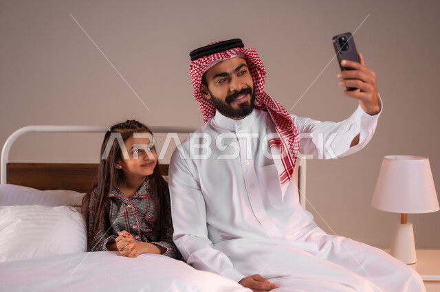 رعاية الأب لأطفاله والعناية بهم، قضاء أوقات ممتعة مع الابناء، مفهوم الأبوة والطفولة، إجراء مكالمة فيديو، بورتريه لرجل عربي خليجي سعودي يجلس على السرير برفقة ابنته يقوم بإلتقاط صورة سيلفي تذكارية بإستخدام الهاتف المحمول، الحب والحنان الاسري، خلفية ملونة
