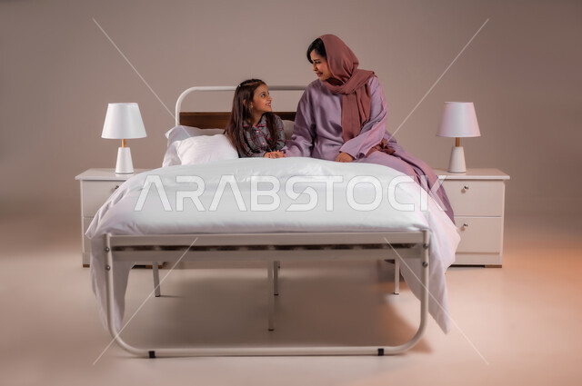 الاسترخاء والاستمتاع بالأجواء الهادئة، مفهوم العناية والاهتمام بالأبناء، قضاء اوقات عائلية سعيدة، بورتريه لامرأة عربية خليجية سعودية محجبة تقوم باجراء محادثة مع ابنتها قبل النوم، اتباع أسلوب حياة صحي، خلفية ملونة