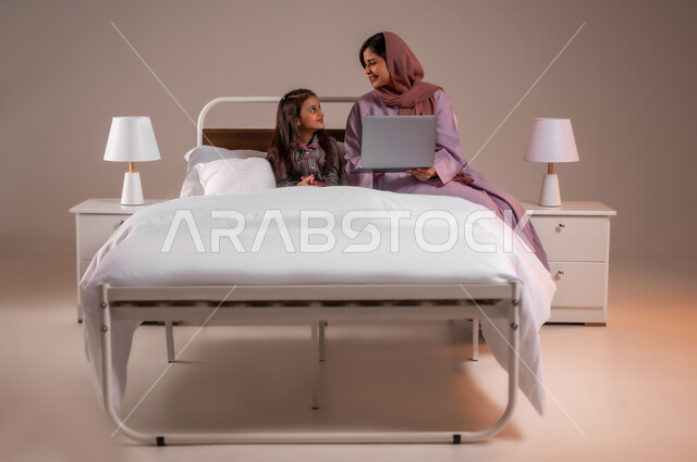 تحمل المسؤولية والعناية بالابناء، متابعة الدروس أونلاين، مفهوم التعلم عن بعد باستخدام الأجهزة التقنية الحديثة والتكنولوجيا، الأسرة السعودية ودورها في تربية إنشاء اجيال المستقبل، بورتريه لامرأة عربية خليجية سعودية محجبة تقوم بمساعدة ابنتها في دروسها، خلفية ملونة